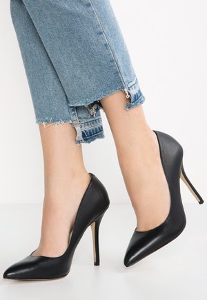 Sorte læder højhælede pumps med spids tå, blank overflade og slank stiletto hæl, parret med slidte blå jeans.