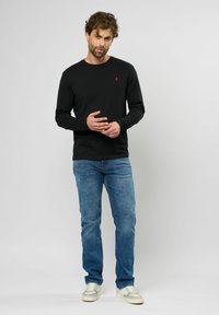 Schwarzes Langarmshirt mit einem kleinen roten Logo, kombiniert mit blauen Jeans und hellen Sneakers. Einfaches Design, Baumwollstoff, lässige Passform.