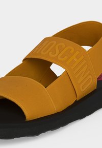 Κίτρινο υφασμάτινο σανδάλι με φαρδιές λωρίδες, "MOSCHINO" ανάγλυφο στη πάνω λωρίδα και μαύρη υφασμάτινη σόλα.