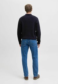 Vīrietis, kurš stāv ar muguru pretī, ģērbts melnā garajā piedurknē un zilās džinsās, kā arī brūnās kurpēs, fona ir vienkārša balta krāsa.