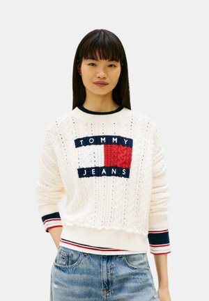 Donna con capelli lisci e scuri che indossa un maglione crema a trecce con il logo "Tommy Jeans" e jeans blu, in piedi davanti a uno sfondo semplice.