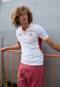 Maillot de football blanc avec des accents rouges et oranges ; logo Puma sur la poitrine. Le modèle porte un pantalon rose. Motif de vagues simple sur le tissu.
