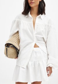 Chemise en lin blanc avec des manches bouffantes, boutonnée à l'avant, associée à une jupe blanche avec taille élastique et texture froncée. Tient un sac en paille.