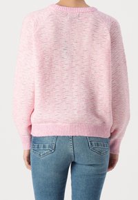 Kobieta w różowym swetrze z fakturowanego dzianiny i niebieskich dżinsach, pokazana od tyłu na jednolitym tle.