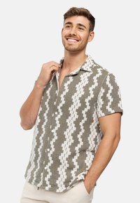 Chemise à manches courtes avec un design à bouton sur le devant qui présente un motif en zigzag en vert olive et blanc. Fabriquée à partir d'un tissu texturé.