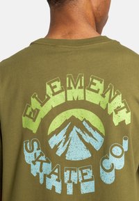 Camiseta de algodón verde oliva con un gran gráfico texturizado y multicolor que presenta montañas y el texto "ELEMENT SKATE CO." en la parte posterior.