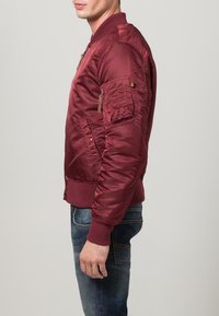 Muž v bordo bomber bundě s kapsou na zip na rukávu a modrých džínách, stojící bokem na šedém pozadí.