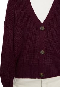 Hluboký fialový hrubý pletený cardigan se třemi velkými hnědými knoflíky, nošený přes bílý top a světlé kalhoty.