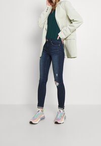 Blazer verde claro, top de rayas verde oscuro, jeans ajustados azul oscuro con detalles rasgados y zapatillas multicolores con cordones.