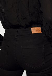 Zwarte denim jeans met een gladde textuur, voorzien van een bruine leren label met de tekst "the blue denim." Slim fit met twee achterzakken.