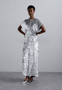 Silver sequin dress with a loose fit, short sleeves, and a long silhouette. Paired with silver pointed-toe shoes on a neutral background.