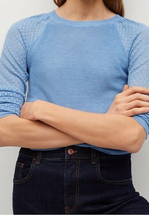 Personne portant un haut en maille bleu clair avec des manches texturées et un jean bleu foncé, les bras croisés à la taille.