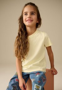 Mladá dívka s dlouhými vlnitými vlasy, oblečená v bledě žlutém tričku a džínech s barevným vzorem záplat, usmívající se a sedící na světle hnědém kostičkovém sedátku.