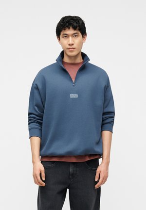 Młody mężczyzna z krótkimi kręconymi włosami, ubrany w niebieską bluzę zapinaną do połowy na zamek, nałożoną na bordową koszulę i czarne dżinsy, stoi na jednolitym tle.