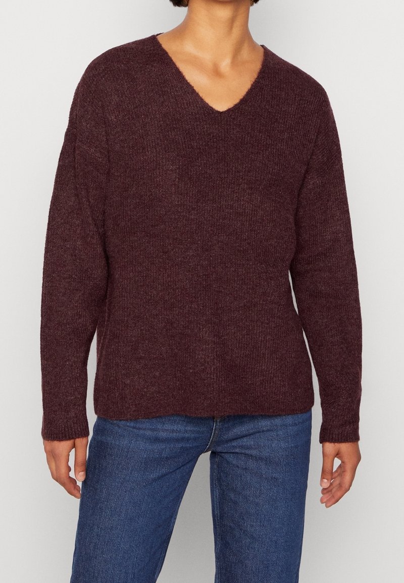 Burgundy stickad tröja med V-ringning, avslappnad passform och långa ärmar, med en mjuk textur och ribbade ärmslut. Matchas med blå jeans.