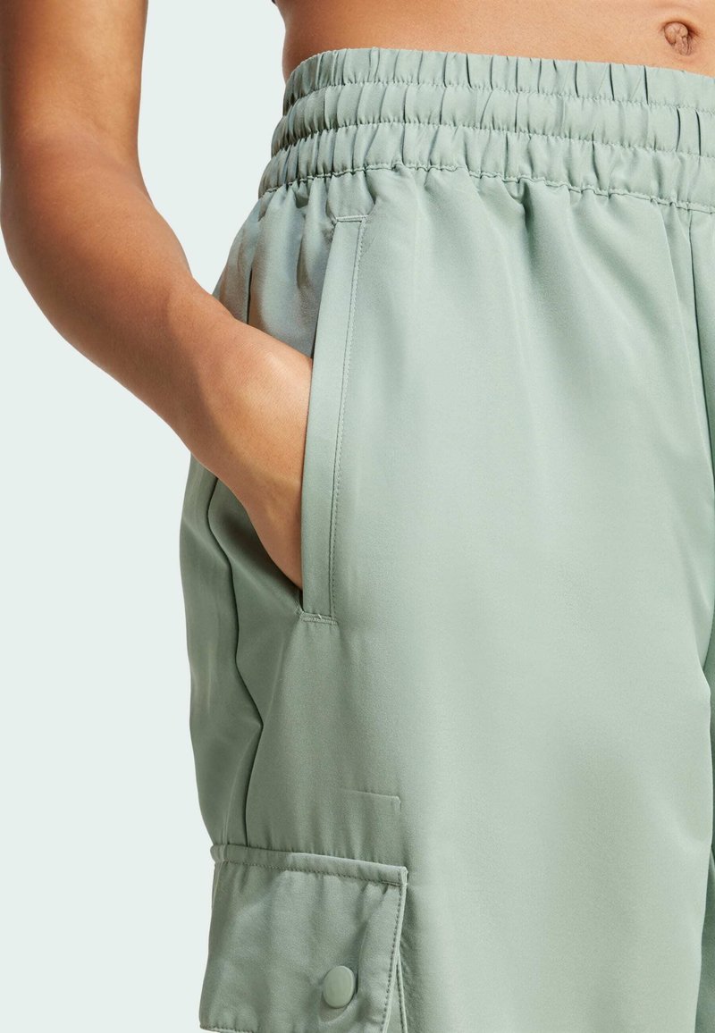 Pantaloni verde chiaro con waistband elastico e tasca laterale. Il tessuto appare liscio con una texture leggermente riflettente.