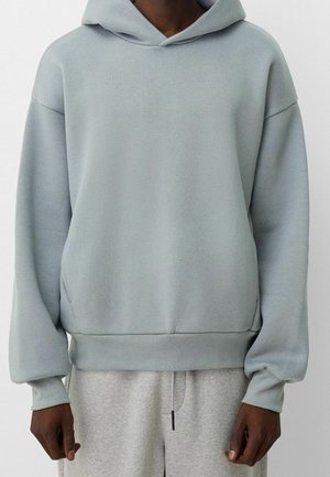 Person iført en lyseblå oversized hættetrøje og lysegrå joggingbukser, stående med armene afslappet ned langs siderne.