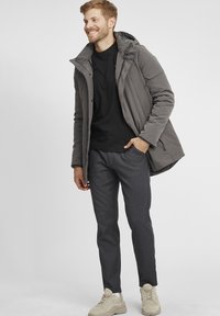 Chaqueta gris aislante con capucha, camiseta negra, pantalones oscuros texturizados y zapatillas de color claro, todo contra un fondo liso.