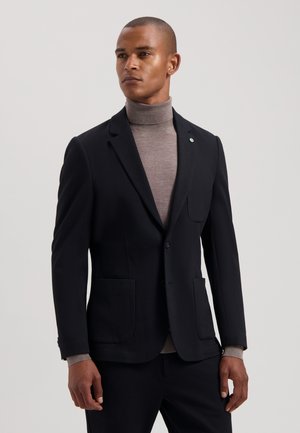 Schwarze Blazer mit strukturierter Oberfläche, Reverskragen, zwei vorderen Taschen und subtilen grünen Akzenten. Gekleidet mit einem hellbraunen Rollkragenpullover.