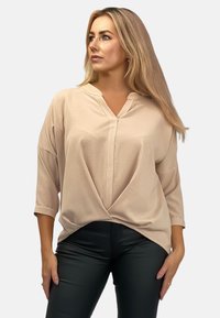 Beżowa, oversize góra z dekoltem w kształcie litery V i rękawami do trzech czwartych. Posiada skręcony przód. Gładki materiał; noszona z czarnymi spodniami.