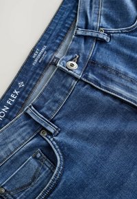 Jeans de mezclilla en azul oscuro con un acabado desgastado. Las características incluyen un cierre de botón, lazos para el cinturón y bolsillos frontales. Etiqueta de la marca visible en la cintura.