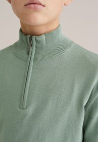 Pull demi-fermeture éclair côtelé vert clair fabriqué en matériau doux, avec un col montant et un détail de fermeture éclair minimaliste à l'avant.