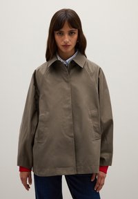 Khaki rain jacket with a pointed collar, two side pockets, and a straight cut. Features red cuffs underneath and smooth texture.