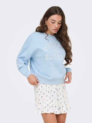Femme ajustant un sweat-shirt bleu clair avec le texte « CHAMPS ÉLYSÉES Paris », portant une jupe blanche fleurie avec un ourlet volanté, debout sur fond blanc.