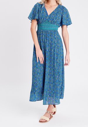 Femme portant une robe midi bleue à motifs avec des manches volantées et une taille smockée, associée à des sandales plates beiges.