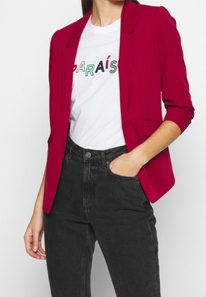 Kobieta w czerwonym blezerze z rękawami 3/4, białym t-shircie z nadrukiem "PARAÍS" i czarnych, wysokim stanem jeansach, z jedną ręką w kieszeni.
