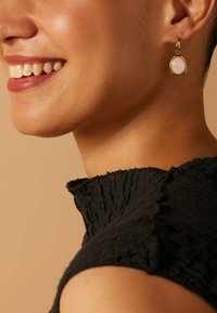 Gouden hangende oorbellen met ovale, witte steencabochons met een gladde afwerking. Het model draagt een gestructureerde zwarte top.