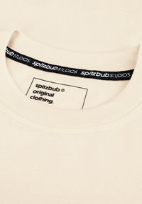 Beige katoenen T-shirt met een brede halslijn, voorzien van een zwart label met witte tekst en een stoffen label dat "spitzbub originele kleding" vermeldt.