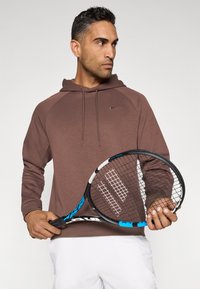Hnedá mikina s kapucňou a raglánovými rukávmi, mäkká textúra a malé logo Nike. Model drží tenisovú raketu s modrým akcentom.