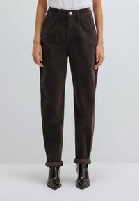 Pantaloni di velluto a coste marroni con vestibilità rilassata, dotati di tasche laterali e risvolti arrotolati, abbinati a stivaletti neri con punta appuntita.