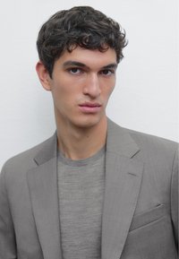 Grau taillierter Blazer über einem hellgrauen Pullover. Schlanke Passform mit Kerbkragen, einer Brusttasche und glatter Textur. Schlichter Hintergrund.