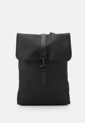 Black minimalist backpack with flap top and buckle strap closure, smooth matte finish, displayed against a plain light background.