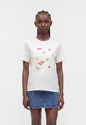 Joven mujer con el pelo trenzado que lleva una camiseta blanca con ilustraciones de comida y una minifalda vaquera azul, de pie frente a un fondo liso.