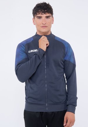Mladý muž v námořnicky modré bundě na zip s modrými vzorovanými rameny, drží zip u brady, stojí na bílém pozadí.