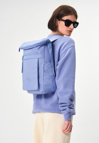 Hellblauer Rucksack mit Klappverschluss, vorderer Reißverschlusstasche und zwei runden Akzenten. Hergestellt aus strapazierfähigem Material mit gepolsterten Trägern.