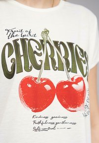 T-shirt en tissu blanc présentant un motif imprimé de deux cerises rouges et un texte stylisé en vert indiquant "FRUIT de l'ESPRIT CERISES".