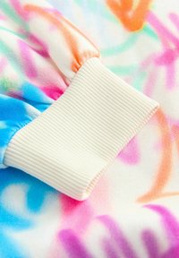 Kolorowa, miękka tkanina ma wzór w wiry w pastelowych odcieniach. Mankiet jest w kolorze kremowym, z prążkowaną teksturą, co podkreśla design odzieży.