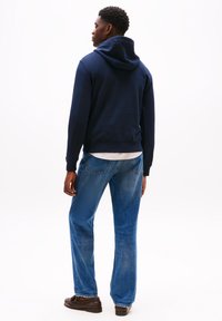 Felpa con cappuccio navy con polsini e orlo a coste, abbinata a jeans in denim blu. I jeans hanno una gamba dritta con tasche posteriori visibili.