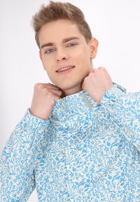 Lichte, met rits sluitende jas in een bloemenpatroon met blauwe en witte kleuren. Heeft een hoge kraag en elastische manchetten. Gladde textuur.