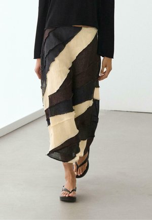 Kvinde går indendørs iført et patchwork midi-nederdel med sorte, beige og brune diagonale stofstriber og sorte thongsandaler.