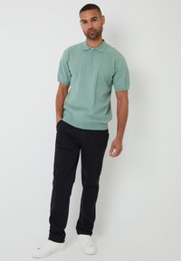 Polo de manga corta en verde menta con un frente texturizado, cierre de cremallera y dobladillo acanalado, combinado con pantalones negros y zapatillas blancas.