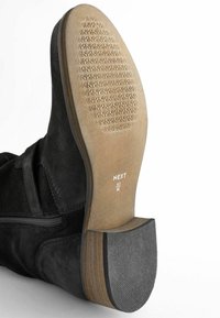 Stivaletto in suede nero con suola in gomma texturizzata, caratterizzato da punta rotonda e cerniera laterale. Il tacco presenta un accento marrone chiaro. Etichetta della taglia visibile.