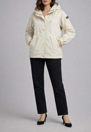 Donna che indossa una giacca con cappuccio color crema con tasche frontali, pantaloni neri e tacchi neri, in piedi contro uno sfondo chiaro e uniforme.