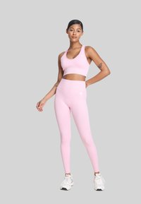 Jasnoróżowy stanik sportowy i legginsy z wysokim stanem, wykonane z gładkiego materiału z detalem logo, zestawione z białymi butami. Modelka stoi na tle szarej ściany.