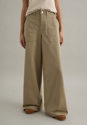 Pantalones anchos color beige con bolsillos parche delanteros, combinados con zapatos de tono marrón claro y una blusa beige claro metida por dentro, sobre un fondo liso.