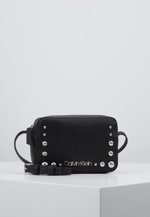Schwarze rechteckige Leder-Umhängetasche mit silbernen Nieten und Calvin Klein-Logo, auf einer weißen Oberfläche vor einem grauen Hintergrund platziert.